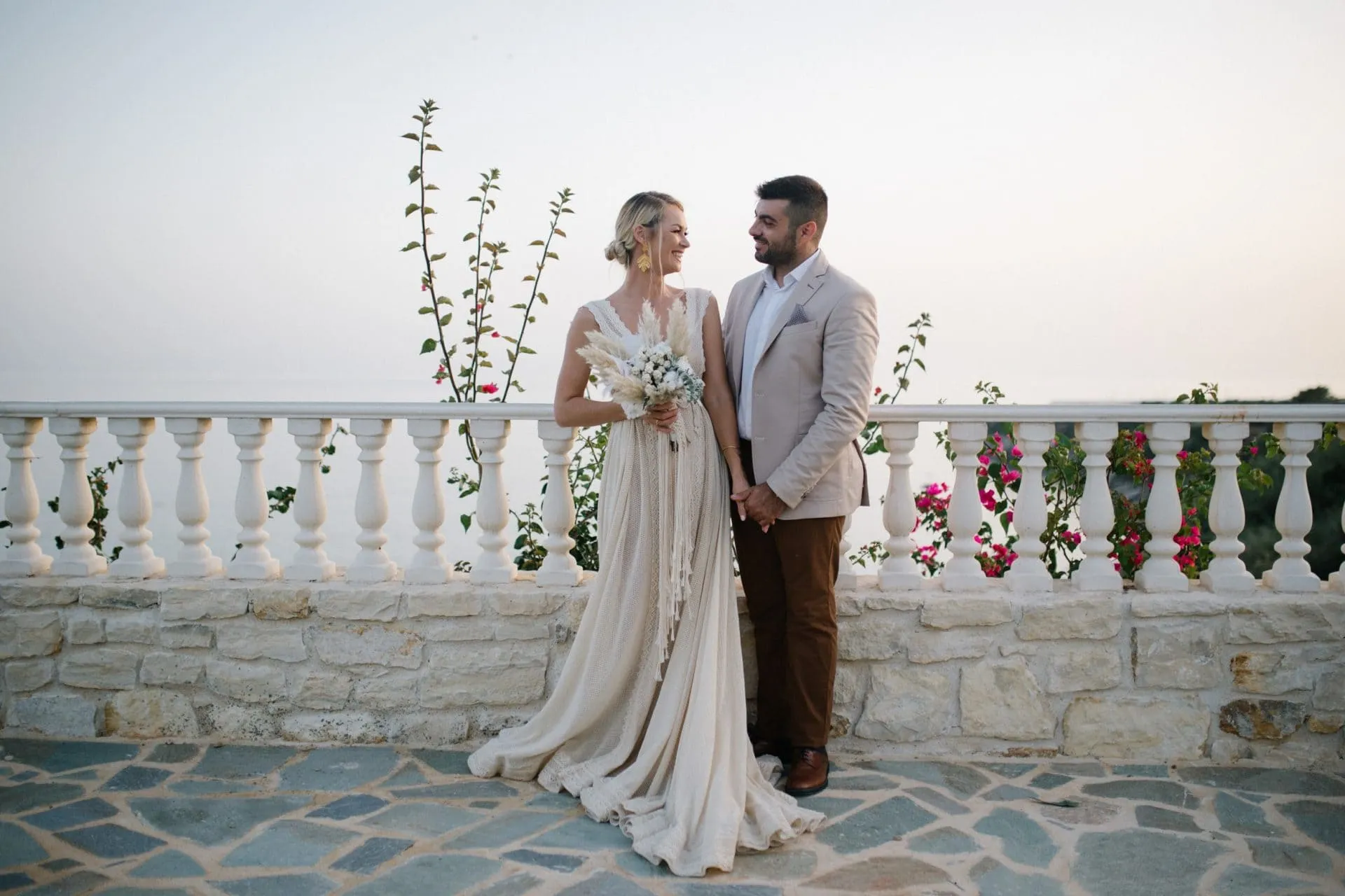 Bride and groom smiling at each other on a flower-lined stone terrace at golden hour